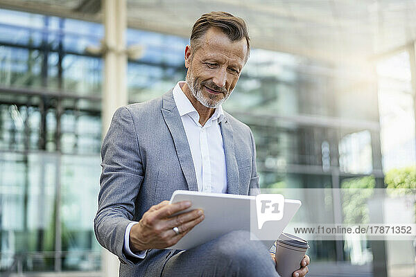 Smiling businessman holding reusable coffee cup using digital tablet