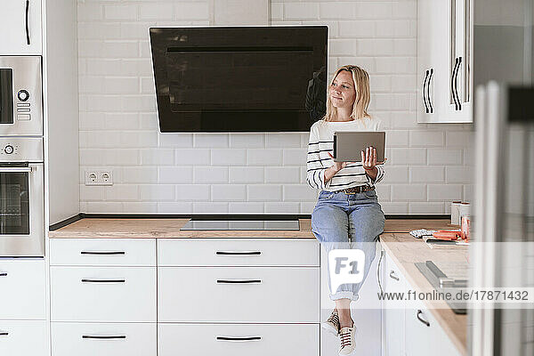 Nachdenkliche Frau mit Tablet-PC sitzt zu Hause auf der Küchentheke