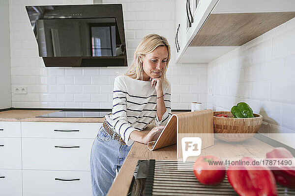 Young woman with hand on chin watching recipe on tablet PC in kitchen at home