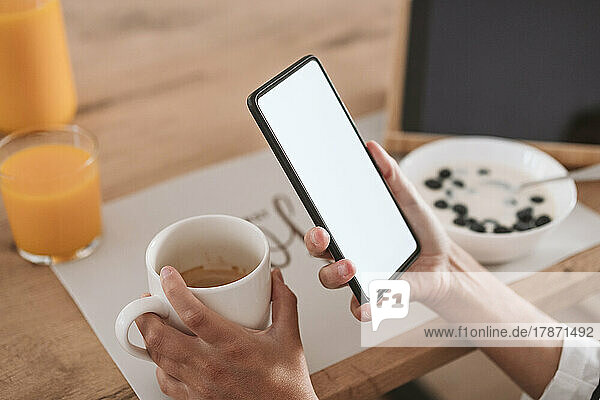 Hands of woman holding coffee cup and smart phone at home