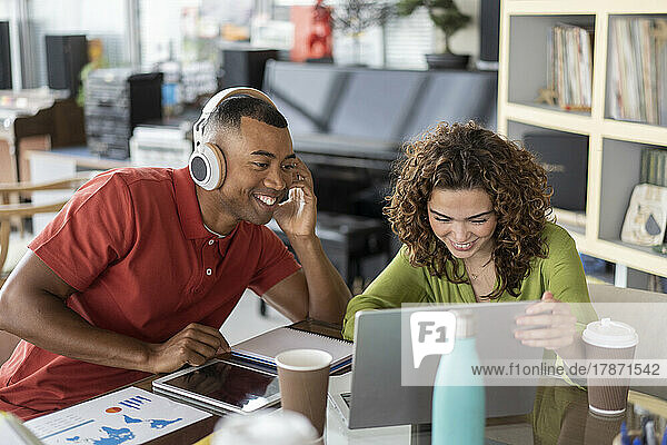 Smiling businesswoman sharing laptop with colleague wearing wireless headphones in office