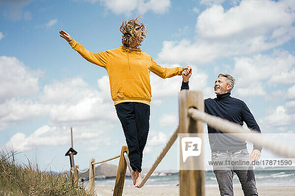 Reifer Mann hält Frau an der Hand und hilft ihr beim Gehen am Seil am Strand