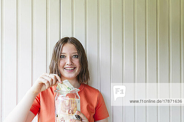 Excited girl saving European currency in jar in front of wall