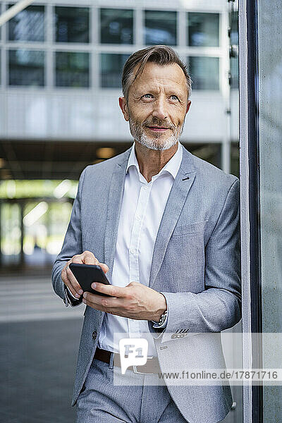 Smiling businessman with smart phone leaning on wall