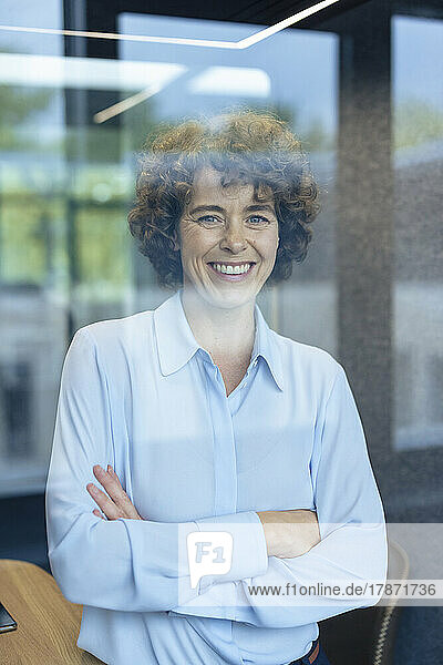 Happy mature businesswoman standing with arms crossed seen through glass