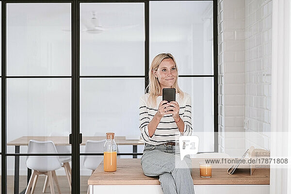 Thoughtful woman with smart phone on kitchen counter at home