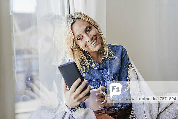 Smiling woman using smart phone leaning on window glass at home