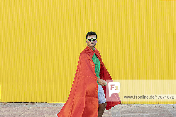 Young man with cape and sunglasses standing in front of yellow wall