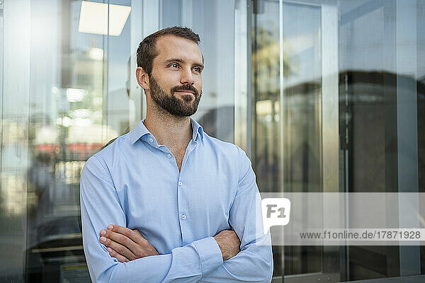 Smiling young businessman with arms crossed standing in front of glass