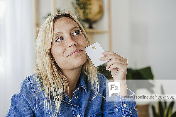 Thoughtful blond woman holding credit card at home