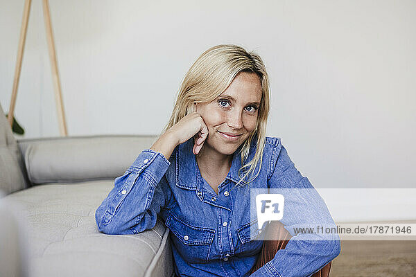 Smiling blond young woman sitting by sofa at home