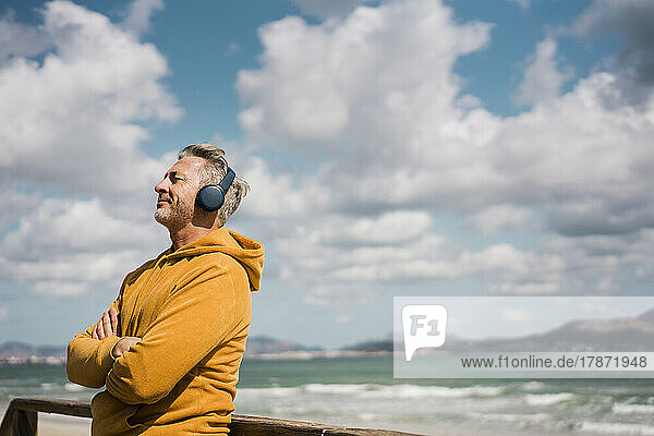 Lächelnder Mann mit kabellosen Kopfhörern steht an einem sonnigen Tag am Strand