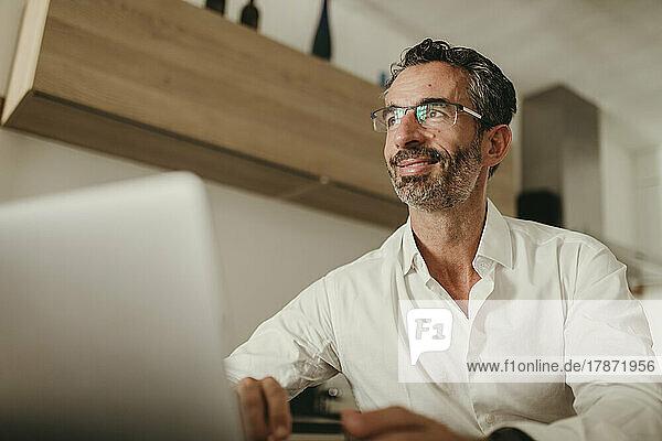 Smiling businessman with laptop contemplating at home