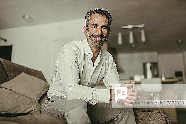 Smiling businessman sitting with hands clasped on sofa at home