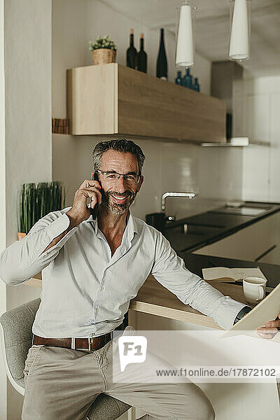 Smiling businessman with document talking on smart phone in kitchen