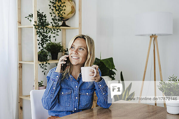 Contemplative businesswoman talking on smart phone holding mug at home office