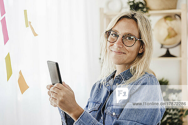 Smiling freelancer with smart phone by adhesive notes at home office