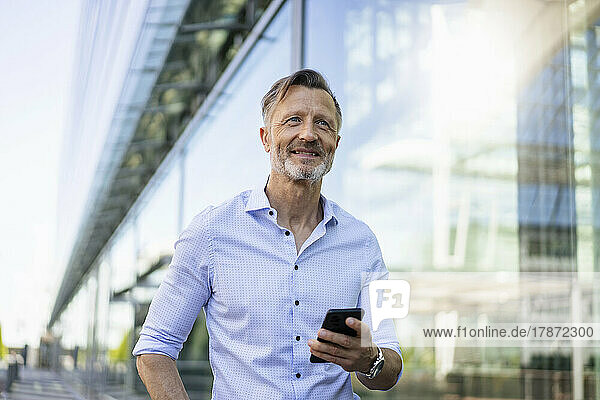 Lächelnder Geschäftsmann mit Smartphone in der Nähe einer Glaswand