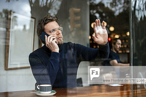 Businessman talking on smart phone touching glass window of cafe