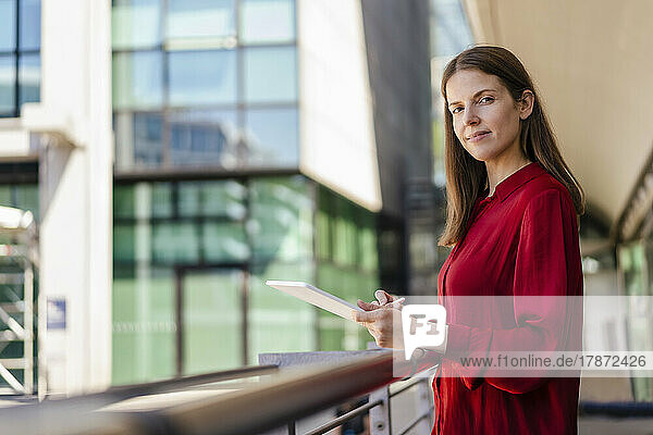 Confident businesswoman with tablet PC by railing at workplace