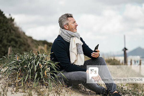 Mature man with smart phone sitting by plant at beach