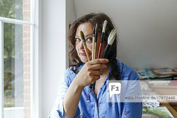 Artist holding paintbrushes by face at home