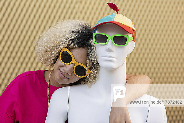 Smiling woman with mannequin wearing sunglasses in front of wall