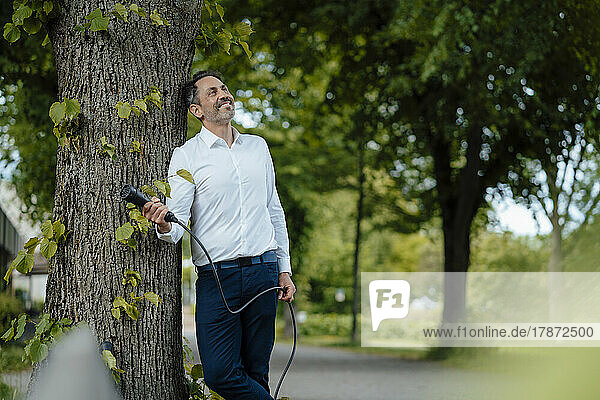 Smiling businessman holding charging cable leaning on tree trunk