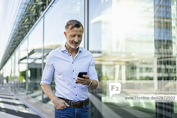 Mature businessman with hand in pocket using smart phone by glass wall