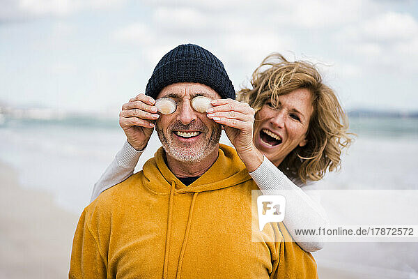 Cheerful woman covering eyes of man with seashells at beach