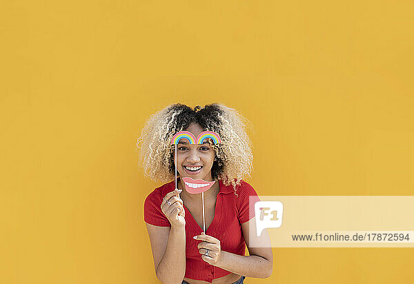 Happy young woman with rainbow and mustache props in front of yellow wall