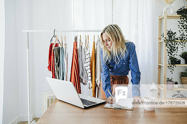 Fashion designer writing on clipboard looking at laptop in home office