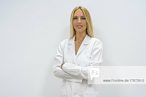 Confident blond doctor with arms crossed standing in front of wall