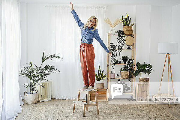 Young woman with arms outstretched standing on stool at home