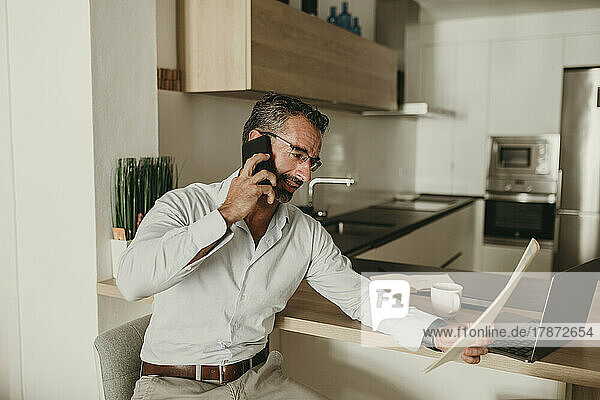 Geschäftsmann liest Dokument und spricht zu Hause in der Küche mit dem Smartphone