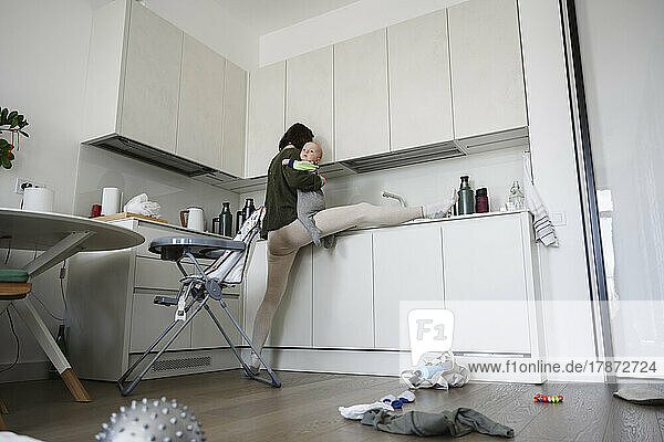 Flexible mother with leg stretched on kitchen counter carrying son at home