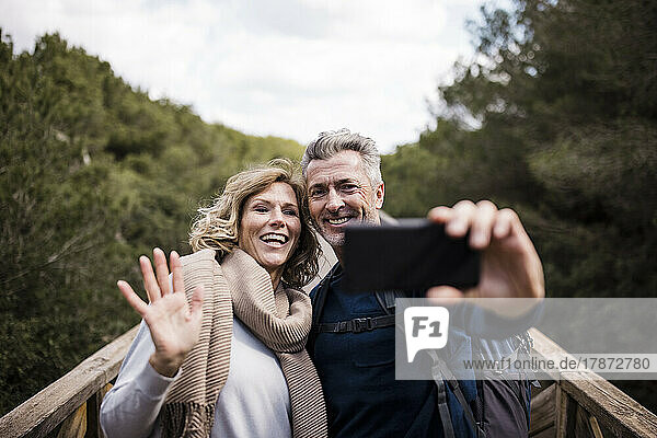 Happy mature man with woman waving hand at smart phone