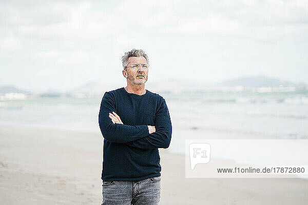 Thoughtful mature man with arms crossed standing at beach