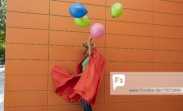 Woman wearing cape holding multi colored balloons in front of wall