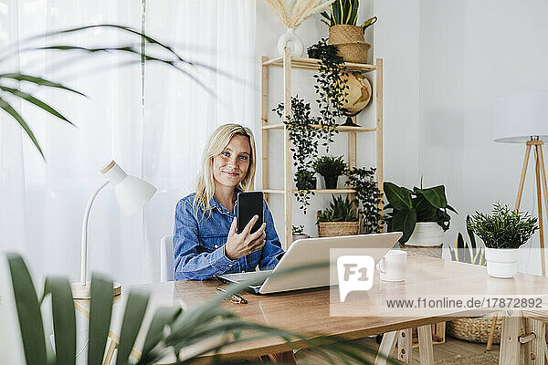 Smiling freelancer with smart phone and laptop sitting at home office