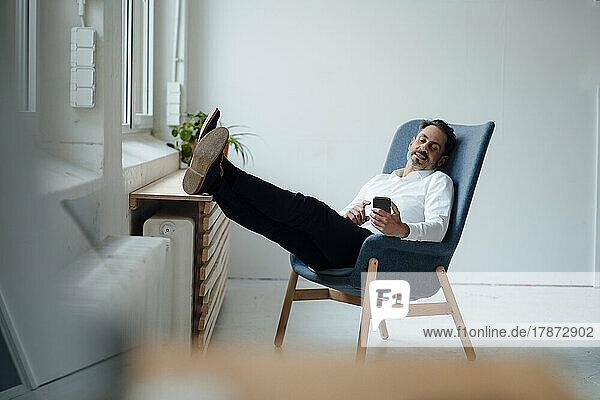 Businessman holding smart phone sitting on armchair at office