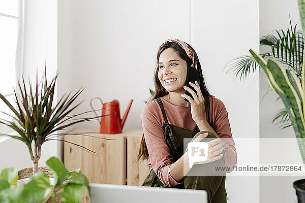 Smiling woman talking on phone sitting by potted plants at home