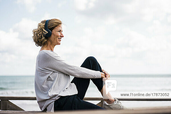 Smiling mature woman wearing wireless headphones sitting at beach