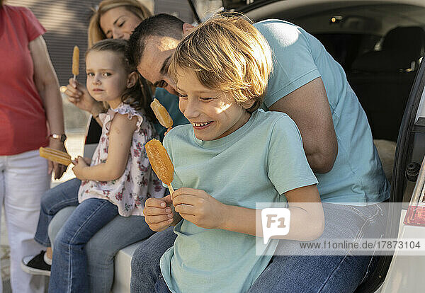Smiling boy with ice pop sitting with family in car trunk