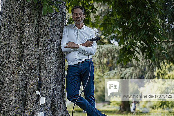Smiling mature businessman with arms crossed holding charging cable leaning on tree trunk