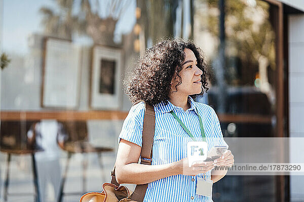 Smiling businesswoman with smart phone standing in front of glass on sunny day