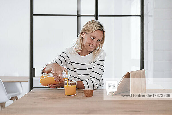 Woman watching tablet PC and pouring juice in glass on table at home