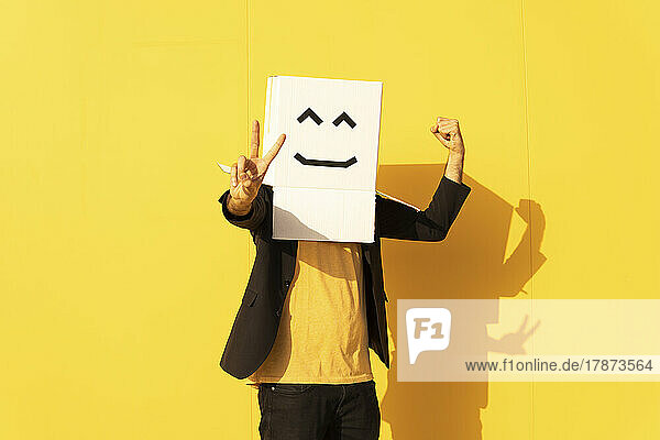 Man wearing box with smiley face gesturing peace sign in front of yellow wall