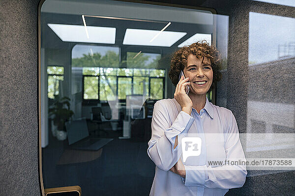 Smiling businesswoman talking on mobile phone leaning on wall seen through glass