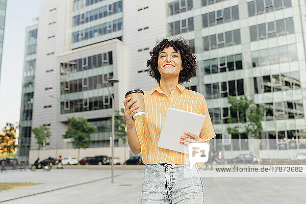 Smiling businesswoman with reusable coffee cup and tablet PC walking in front of building